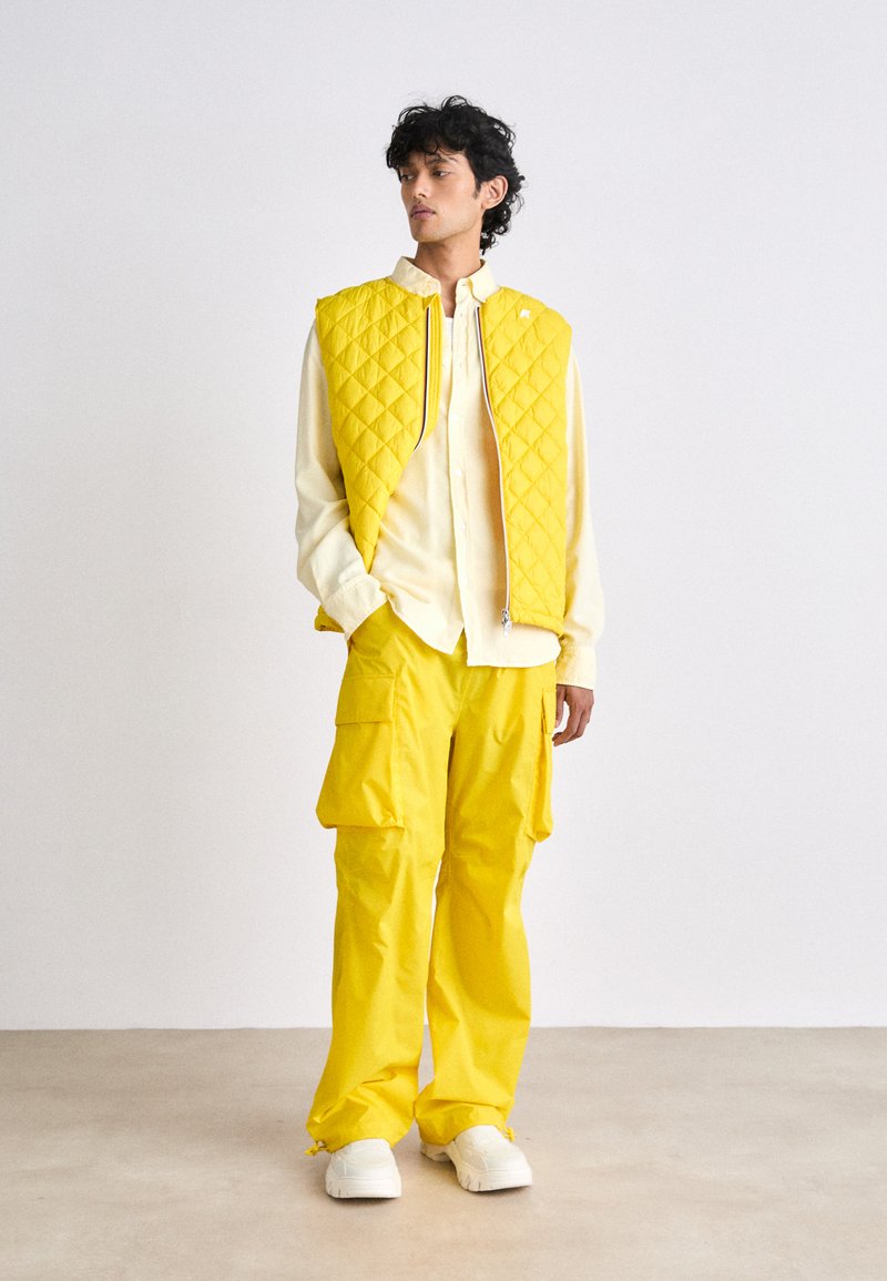 Gilet matelassé jaune associé à une chemise boutonnée jaune clair et un pantalon cargo assorti. Il comprend une fermeture éclair et des poches spacieuses.