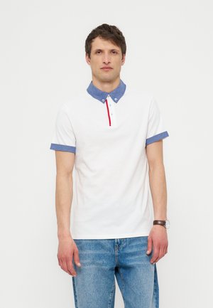 Mężczyzna w białej koszulce polo z niebieskim kołnierzykiem i mankietami, niebieskich dżinsach i zegarkiem na rękę, stojący na jednolitym jasnym tle.