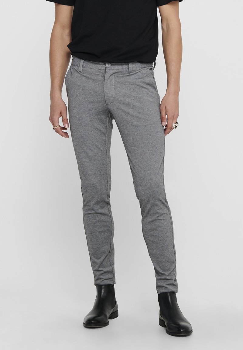 Pantalones gris ajustados de tela texturizada, con cierre de cremallera, botón y bolsillos laterales, combinados con botines negros de tobillo.