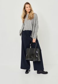 Graue Strickjacke, marineblaue weite Hose, schwarze Lackledertasche mit Nietenakzenten und schwarze Schnürstiefel. Modell steht vor einem hellen Hintergrund.