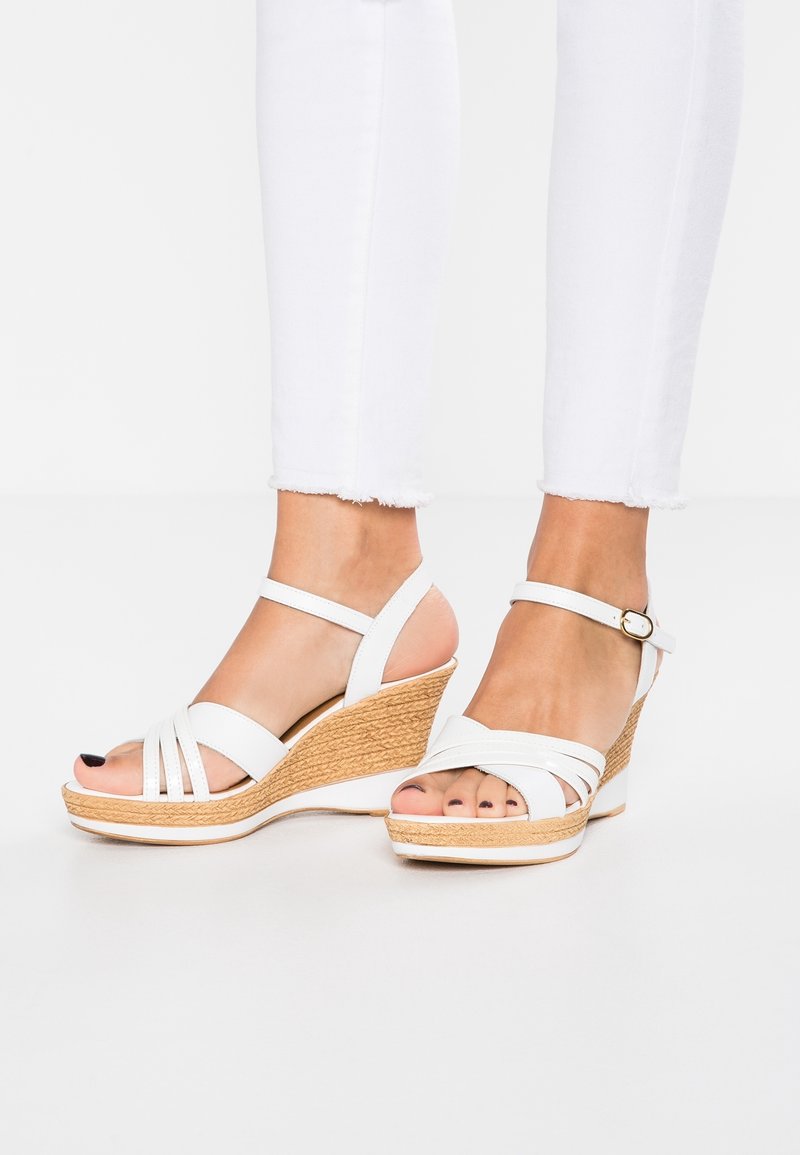 Pieds portant des sandales compensées blanches avec des brides à la cheville et des talons tressés texturés, associées à un pantalon blanc court sur un fond uni.