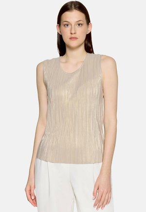 Frau mit glatten braunen Haaren, die ein ärmelloses, beiges, plissiertes Oberteil und weiße Hose trägt und vor einem schlichten hellen Hintergrund steht.