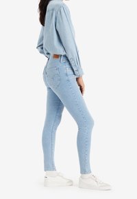 Jeans ajustados en denim azul claro, con tiro alto, diseño de cinco bolsillos y distintivos bolsillos traseros en forma de corazón. Combinados con zapatillas blancas.
