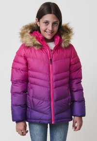Chica con una chaqueta de invierno de degradado de rosa a morado y capucha forrada de pelo, sonriendo, de pie frente a un fondo liso.