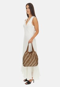 Borsa intrecciata marrone e crema con due manici marroni. La borsa ha una forma morbida e una superficie texturizzata. Il modello indossa un lungo vestito bianco e tacchi neri.