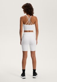 Bijeli sportski crop top s dizajnom na leđima u obliku križa, uparen s bijelim šortsem do koljena. Glatka, rastezljiva tkanina. Crna obuća i čarape.