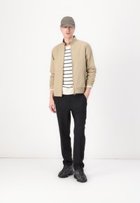 Beige lichtgewicht jack met een opstaande kraag, witte en zwarte gestreepte trui, donkere broek en zwarte met patroon sneakers. Olijfkleurige pet.