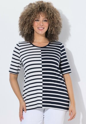 Frau mit lockigem Haar, die ein kurzärmeliges Oberteil mit schwarz-weiß gestreiftem Muster und weiße Hosen trägt, lächelt vor einfarbigem Hintergrund.