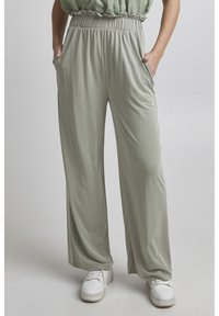 Light green wide-leg pants made of a smooth, stretchy fabric, featuring an elastic waistband and side pockets, paired with white sneakers.