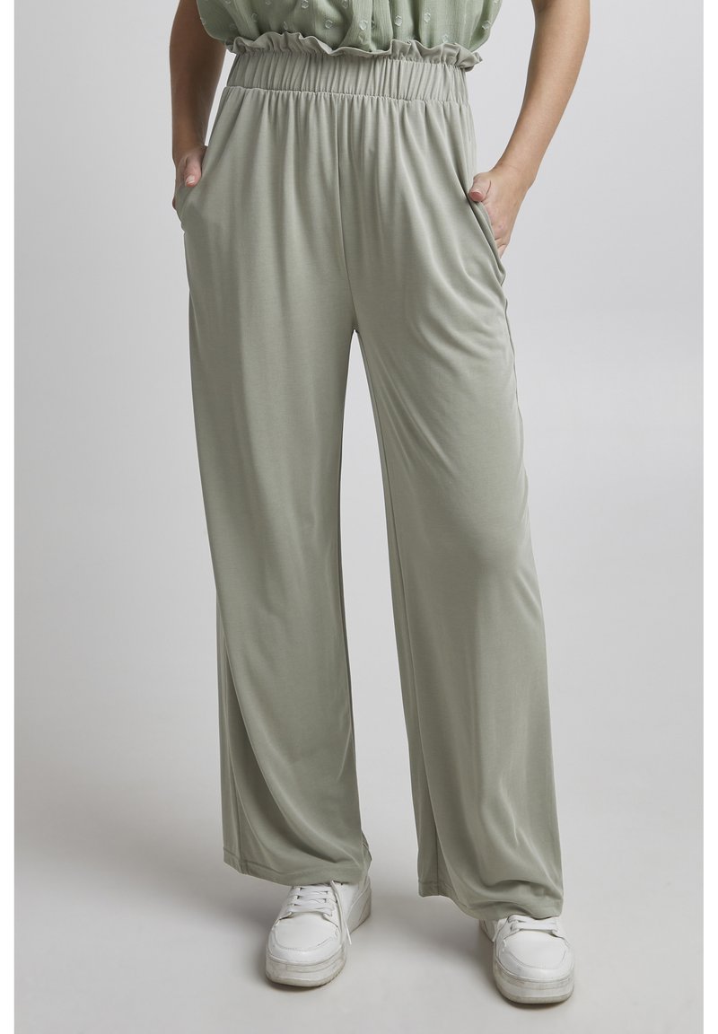 Light green wide-leg pants made of a smooth, stretchy fabric, featuring an elastic waistband and side pockets, paired with white sneakers.