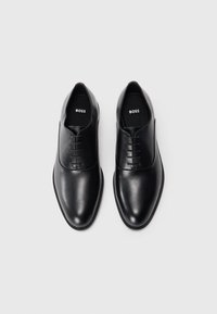 Une paire de chaussures formelles en cuir noir à lacets avec des orteils ronds et un logo "BOSS" sur les semelles intérieures, photographiée sur fond blanc.