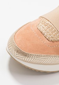 Persikofärgad mockasneaker med ett beige elastiskt band, texturerad vävd detalj och en ljus gummisula. Sömorna synliga längs kanterna.