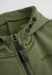 Oliwkowa bluza z kapturem na zamek, z czarnymi elementami zamka, kołnierzem wykończonym polarem oraz nadrukowanym tekstem na materiale.