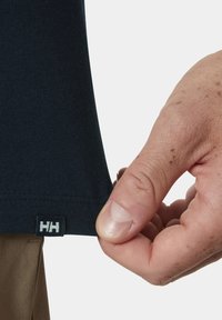 Camiseta de algodón azul marino, con mangas cortas y una pequeña etiqueta con el logo "HH" cerca del dobladillo, sostenida por una mano.