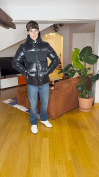 Geacă puffer neagră cu un finisaj neted și un panou mat contrastant, purtată cu blugi albaștri și adidași albi, stând în interior.