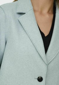 Cappotto in misto lana blu chiaro con colletto a punta e chiusura a singolo bottone nero. Texture liscia, vestibilità sartoriale e dettagli di cucitura sottili.