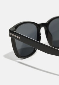 Occhiali da sole neri con una cornice rettangolare audace, lenti grigie e dettagli metallici sulle tempie. Texture liscia e design elegante.