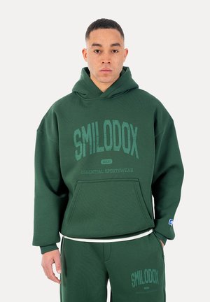 Mężczyzna ubrany w ciemnozieloną bluzę z kapturem i dopasowane spodnie dresowe z logo „SMILODOX Essential Sportswear” na obu elementach odzieży.