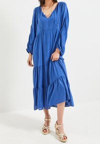 Modrá, dlouhá šatová šaty s vrstvami vyrobené z lehkého materiálu, s výstřihem do V a sklady; doplněné sandály na podpatku s pásky.