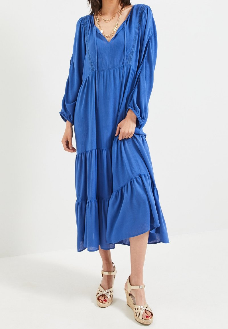 Modrá, dlouhá šatová šaty s vrstvami vyrobené z lehkého materiálu, s výstřihem do V a sklady; doplněné sandály na podpatku s pásky.