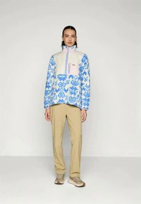 Pull en polaire avec un motif floral bleu et un panneau crème ; il présente un col montant, une fermeture éclair et des touches de rose. Associé à un pantalon beige clair.