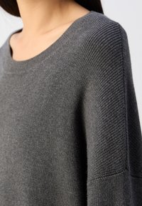 Femme portant un pull en maille côtelée gris foncé avec un col rond, montré de l'épaule à la partie supérieure de la poitrine.