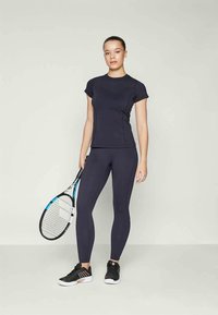 Top atlético azul-marinho e leggings feitas de tecido elástico. Modelo segura uma raquete de ténis, vestindo sapatos pretos com solas brancas.