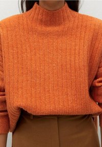 Pull à col roulé côtelé en orange vif, fait de tissu texturé. Coupe ample avec épaules tombantes, associé à un pantalon marron.