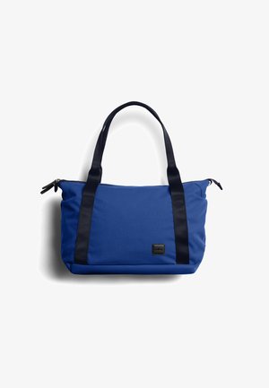 Sac cabas bleu avec poignées noires et fermeture éclair, écusson logo rectangulaire à l'avant, fond uni.