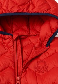 Chaqueta acolchada roja con capucha, con un ribete azul marino. Cierre de cremallera con un parche de logo en el pecho. Textura suave y acolchada.