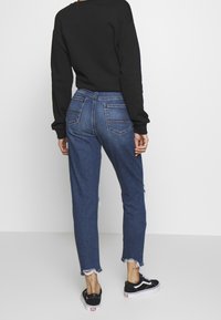 Högmidjade blå denimjeans med slitna fållar, med bakfickor och en avslappnad passform, kombinerat med svarta sneakers.