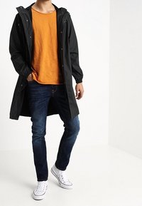 Svart vattentålig jacka med dragsko i huvan, orange T-shirt, blå jeans och vita sneakers. Har tryckknappar och elastiska manchetter.