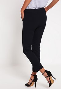 Osoba oblečená v upnutých černých kalhotách a černých vysokých podpatcích stojí s rukama v zadních kapsách na bílém pozadí.