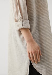 Chemise à carreaux léger beige en tissu transparent ; possède des manches retroussées et une fente sur le côté, montrant une surface texturée.