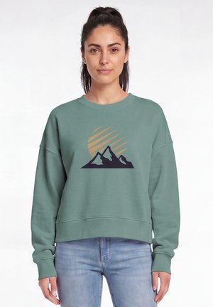 Femme aux cheveux foncés attachés, portant un sweat-shirt vert avec un motif montagne et soleil et un jean bleu clair, debout devant un fond blanc.