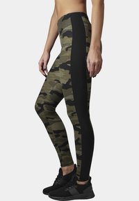 Camo-patterned leggings featuring a blend of green hues with black side panels; made of stretch fabric. Black sneakers complete the look.