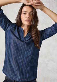 Top in denim blu scuro con righe bianche verticali, caratterizzato da scollo a V e maniche tre quarti arricciate. Tessuto liscio e vestibilità morbida.
