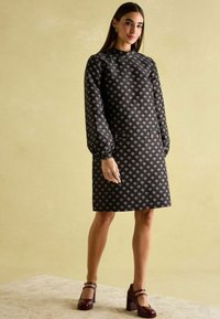 Schwarzes Kleid mit langen, gepufften Ärmeln und hohem Kragen, das ein sich wiederholendes florales Muster in Rosa und Creme zeigt. Kombiniert mit burgunderfarbenden High Heels.