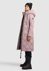 Cappotto lungo imbottito rosa chiaro con trapuntatura a diamante, cappuccio e zip nera. Abbinato a stivaletti da combattimento neri con lacci e un berretto.