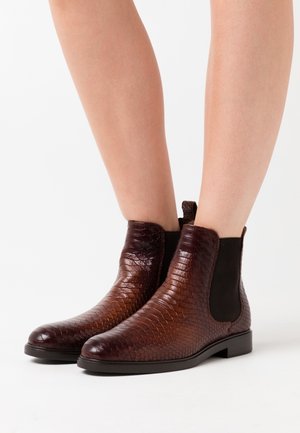 Brown textured leather ankle boots with elastic side panels worn on bare legs against a white background.