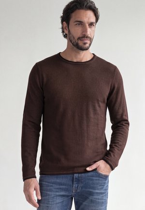 Mann mit dunklem Haar und Bart trägt ein braunes Langarmhemd und blaue Jeans, steht mit einer Hand in der Tasche vor einfachem Hintergrund.