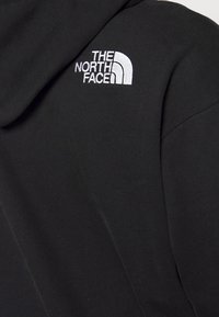 Svart huvtröja med en vit broderad logotyp av "The North Face" på vänster axel, tillverkad av mjukt bomullstyg.