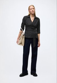 Zwarte blouse met witte stippen, wikkelontwerp en driekwart mouwen, gecombineerd met donkerblauwe jeans met wijde pijpen en een lichtbeige tas.