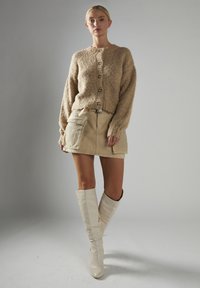 Braunfarbige, strukturiert gestrickte Cardigan mit Knöpfen, beiger Cargo-Rock mit Taschen und weißen kniehohen Stiefeln. Minimalistisches Design und neutrale Farben.