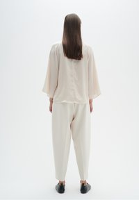 Blusa leggera di colore crema con maniche ampie, abbinata a pantaloni larghi di colore crema e scarpe nere, vista di spalle.