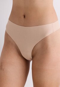 Nahaufnahme einer Person, die nahtlose hautfarbene Unterwäsche trägt, wobei der untere Bauch und die Oberschenkel vor einem schlichten Hintergrund zu sehen sind.