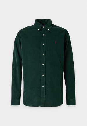 Jeune homme debout portant une chemise boutonnée vert foncé, un jean large bleu clair et des chaussures beiges sans lacets sur un fond blanc uni.