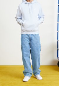 Ljusblå hoodie med framficka och vit Nike-logga, ihop med lössittande blå jeans och vita sneakers, mot en gul bakgrund.