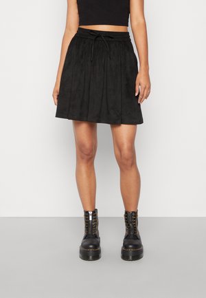 Femme portant un crop top noir, une jupe taille haute noire avec un cordon et des bottines à plateforme noires lacées, debout sur un sol gris.