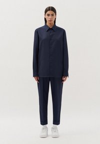 Camicia button-up blu navy con colletto aperto, abbinata a pantaloni tapered coordinati. Indossata con sneaker bianche, il tessuto appare liscio e leggero.
